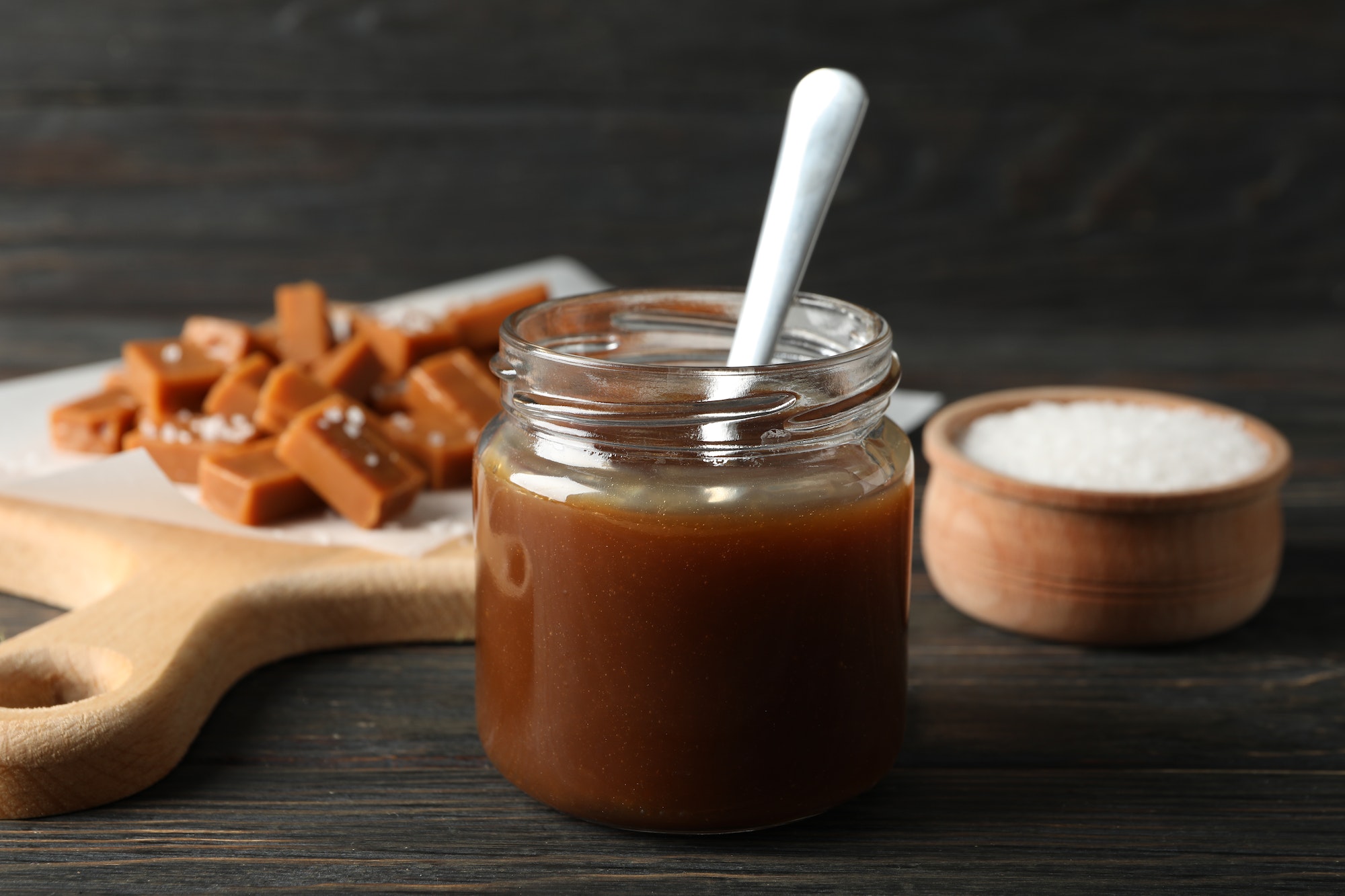 Glass jar with salted caramel and candies on wooden background, space for text